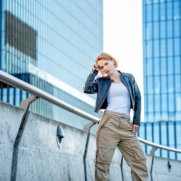 Dorota Stokowiec Fotografia - Kobieta w skórzanej kurtce i spodniach cargo pozuje na tle nowoczesnej architektury. Uwagę przyciągają detale stroju i miejski krajobraz w tle, tworząc modny, dynamiczny portret.