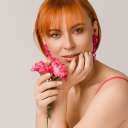 Dorota Stokowiec Fotografia - Portret kobiety z rudymi włosami, w różowej sukience i kolczykach, trzymającej bukiecik różowych goździków przy twarzy, na jasnym tle.
