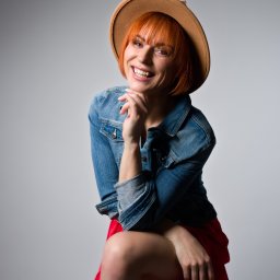 Dorota Stokowiec Fotografia - Uśmiechnięta, ruda kobieta w dżinsowej kurtce i kapeluszu, siedząca na krześle na szarym tle. Jasne oświetlenie podkreśla detale stroju i wyraz twarzy.