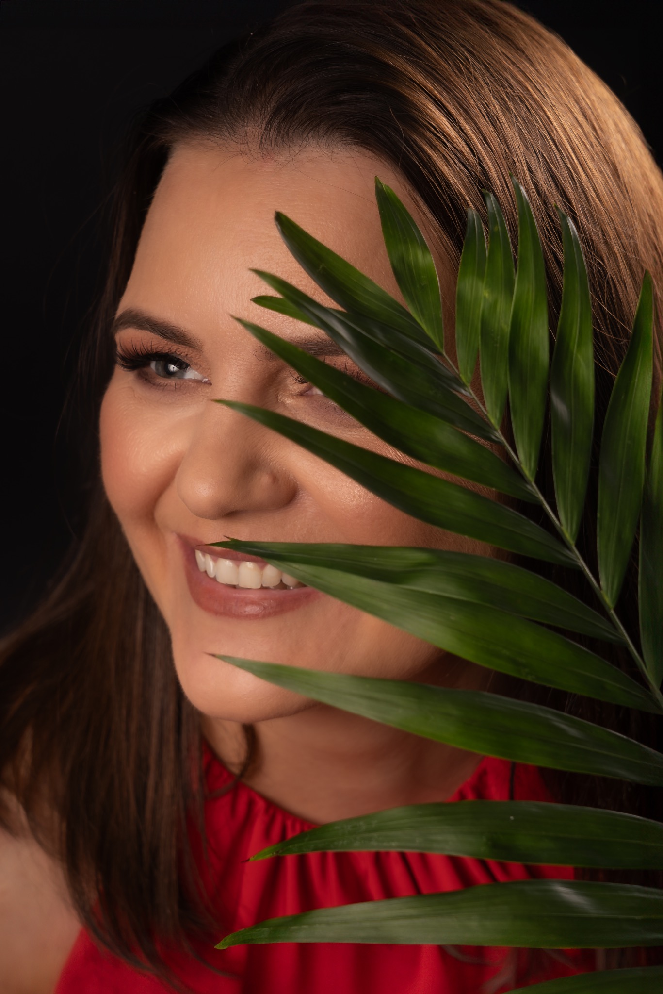 Portret uśmiechniętej kobiety z długimi brązowymi włosami, częściowo zasłoniętej liściem palmy na czarnym tle, w czerwonej sukience.