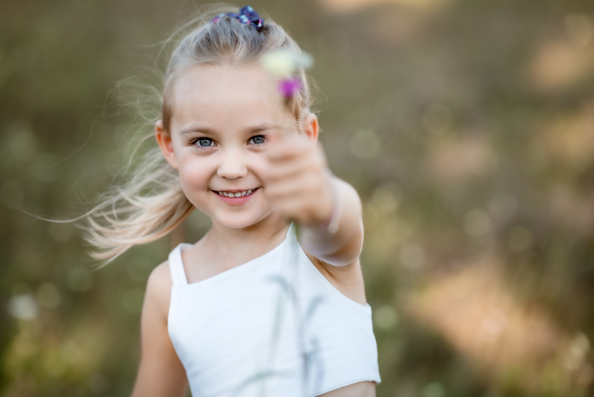 Uśmiechnięta blond dziewczynka z niebieskimi oczami trzyma rozmazany kwiat na wyciągniętej ręce, w tle zieleń.