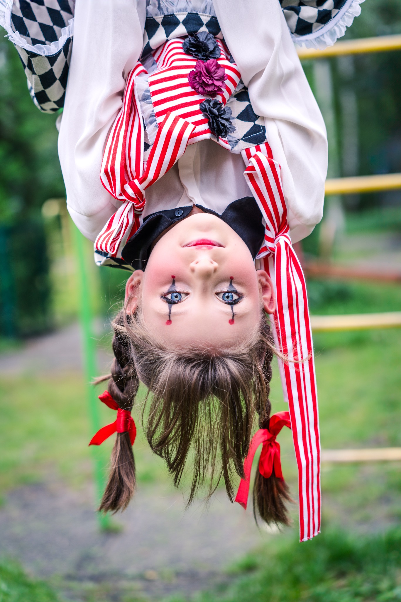 Dziewczynka w kostiumie klauna z pomalowaną twarzą, wisząca do góry nogami na tle zieleni.