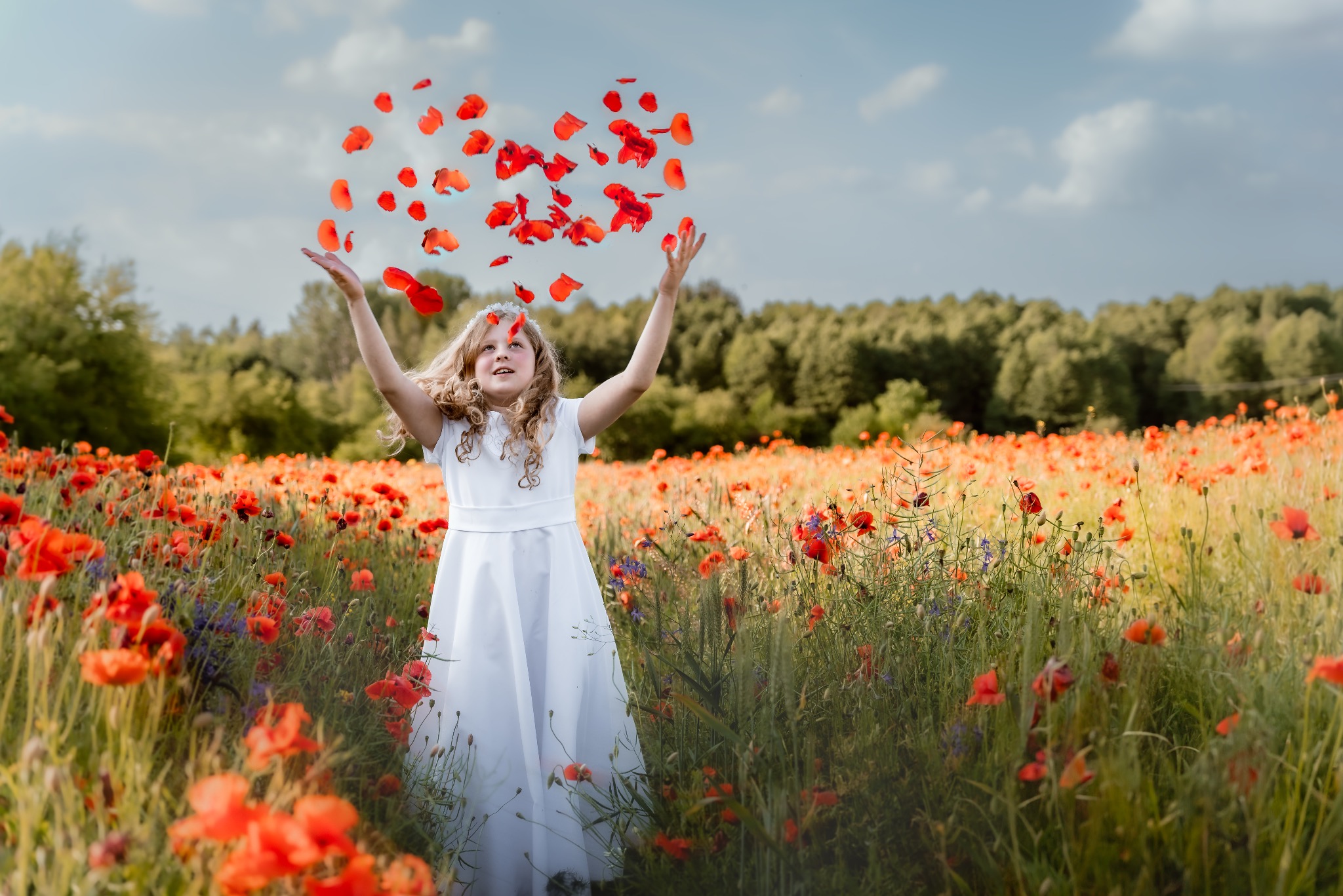 Dziewczynka w białej sukience i wianku na głowie, stojąca na łące maków, podrzuca w górę płatki kwiatów.