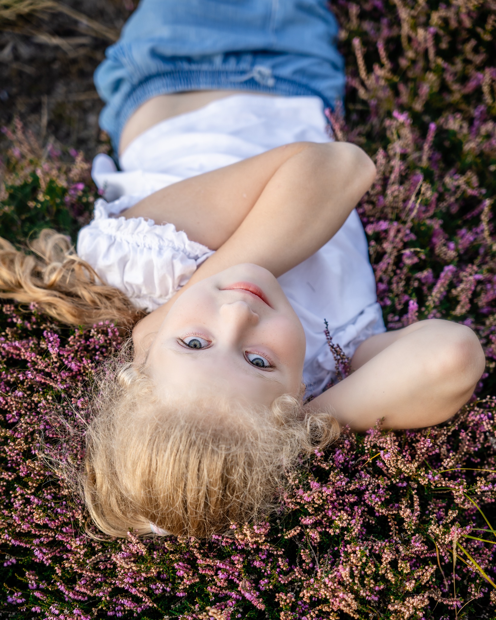 Dziewczynka z długimi blond włosami leży na plecach w kwitnącym wrzosowisku, ubrana w białą bluzkę i dżinsowe spodenki, patrzy w górę.