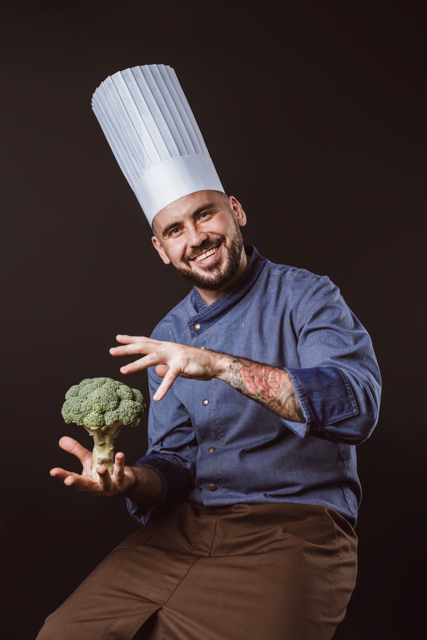 Uśmiechnięty kucharz w wysokiej czapce trzyma brokuła w dłoni, prezentując go na ciemnym tle.