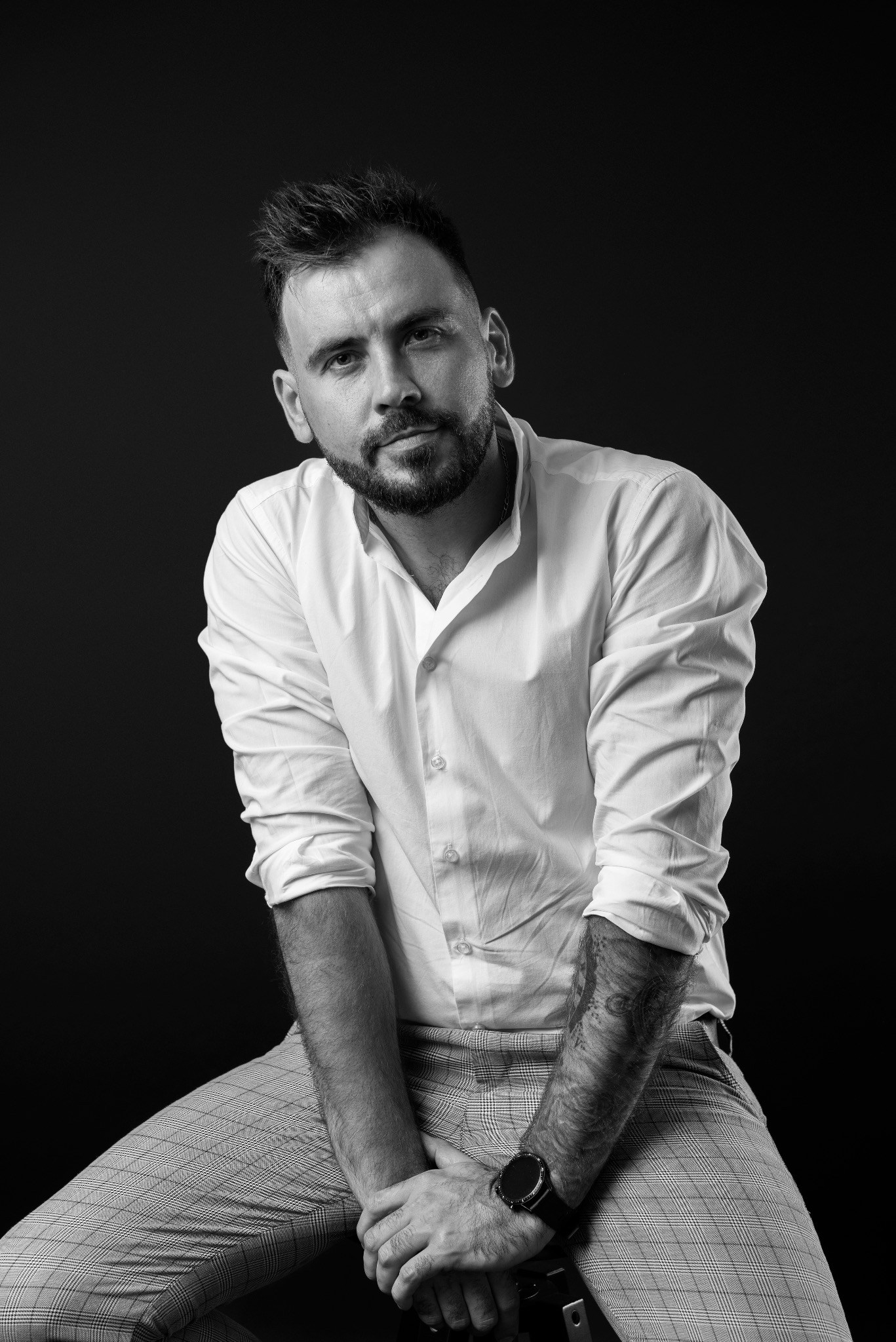 Czarno-białe portretowe ujęcie mężczyzny z brodą, w białej koszuli i spodniach w kratę, siedzącego na krześle na czarnym tle, z widocznym tatuażem na przedramieniu i zegarkiem na nadgarstku.
