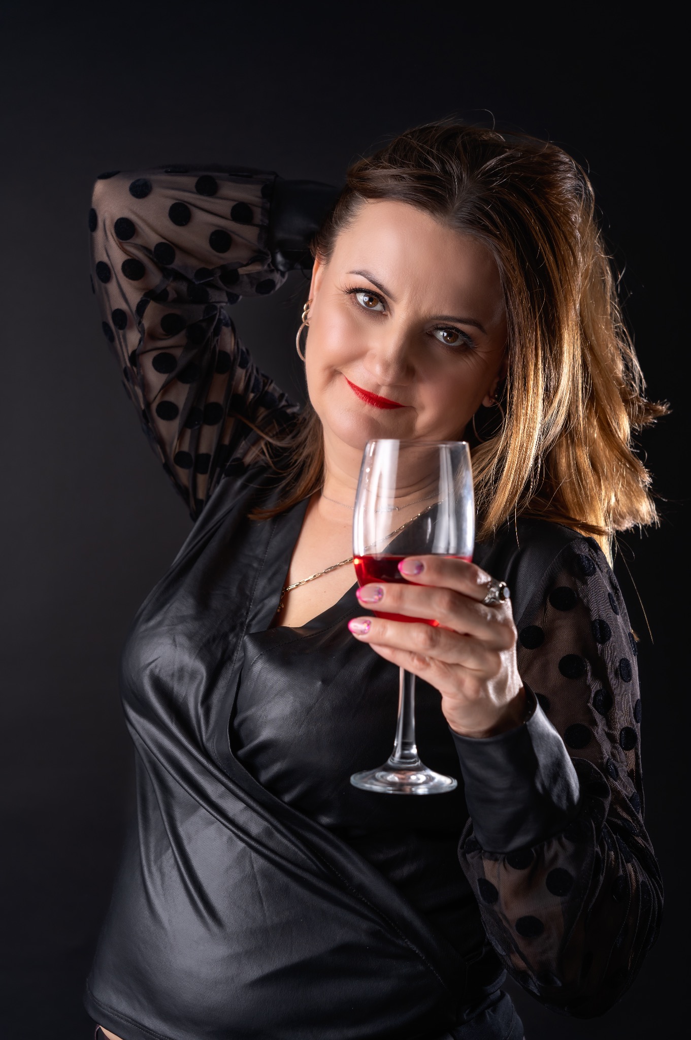 Kobieta w czarnej bluzce w kropki, z czerwonym winem w kieliszku, uśmiecha się do kamery na ciemnym tle.