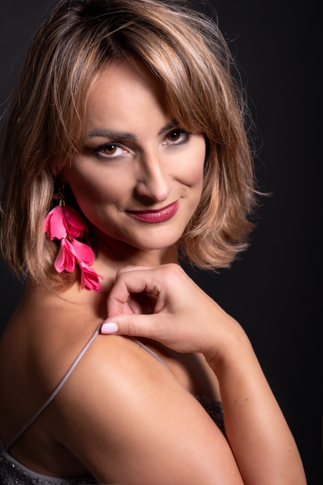 Portret kobiety z krótkimi blond włosami, w różowych kolczykach w kształcie kwiatów, delikatnie dotykającej ramiączka sukienki na czarnym tle.