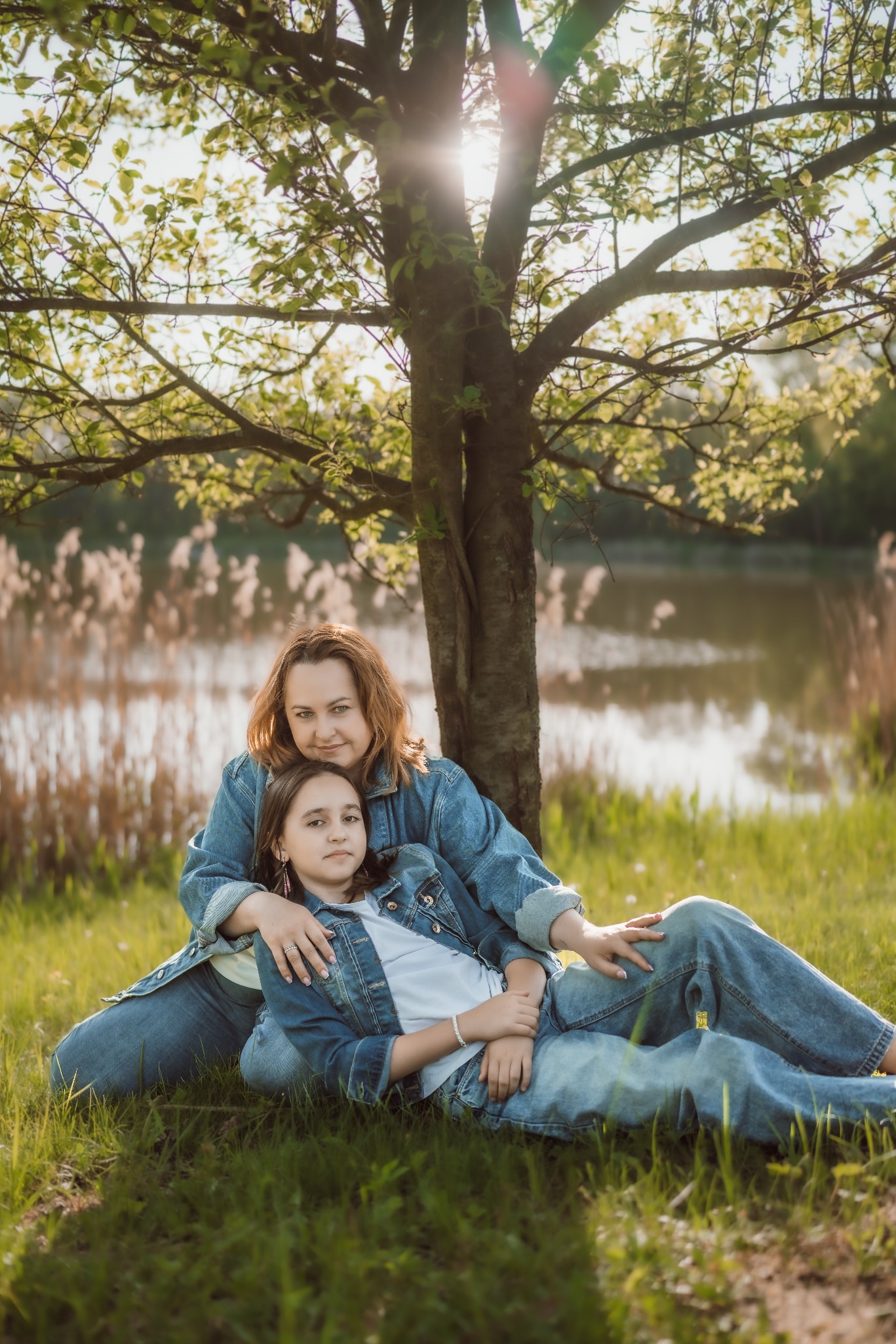 Mama i córka w dżinsowych kurtkach pozują na trawie pod drzewem w słoneczny dzień nad brzegiem jeziora. Portret w naturalnej scenerii.