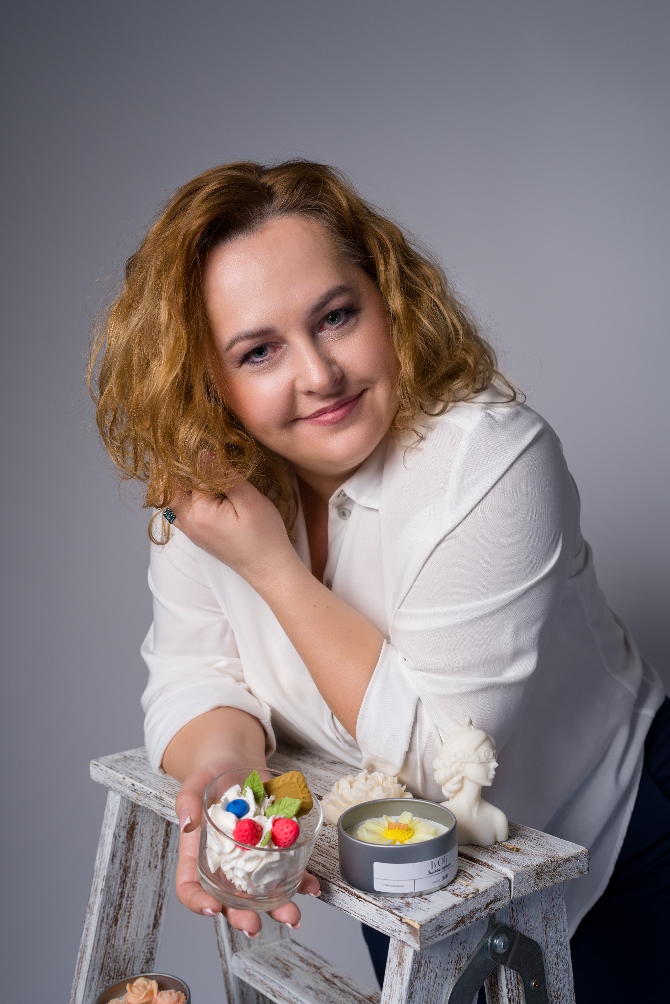 Portret kobiety w białej koszuli, opierającej się o białą, drewnianą drabinkę, na której znajdują się dekoracyjne świece i deser w szklance na szarym tle.