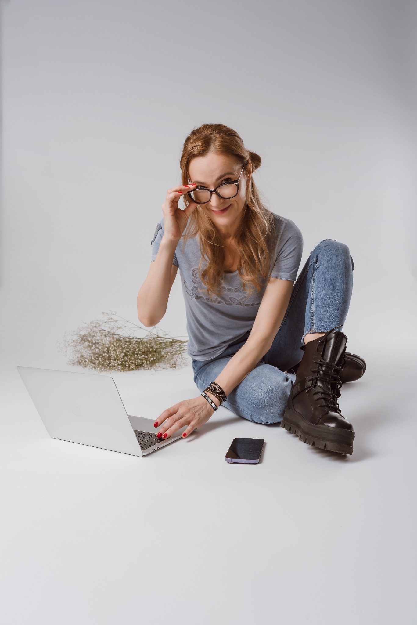 Kobieta w okularach siedzi na podłodze z laptopem, telefonem i kwiatami. Uśmiecha się, dotykając oprawki okularów. Stonowana kolorystyka, jednolite tło.