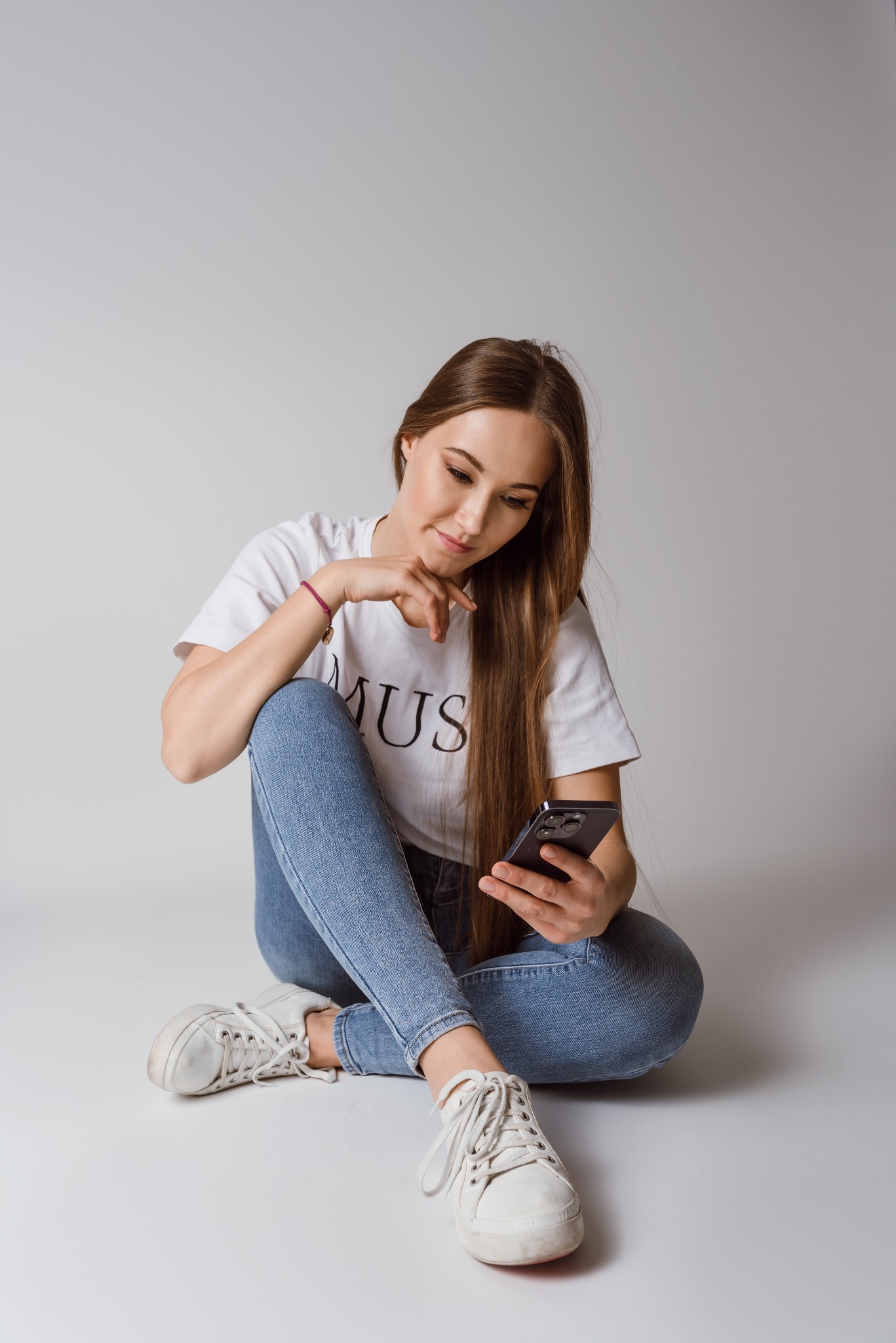 Młoda kobieta z długimi włosami, w dżinsach i białej koszulce z napisem, siedzi na podłodze i patrzy na smartfon w ręku, uśmiechając się lekko. Białe tło.