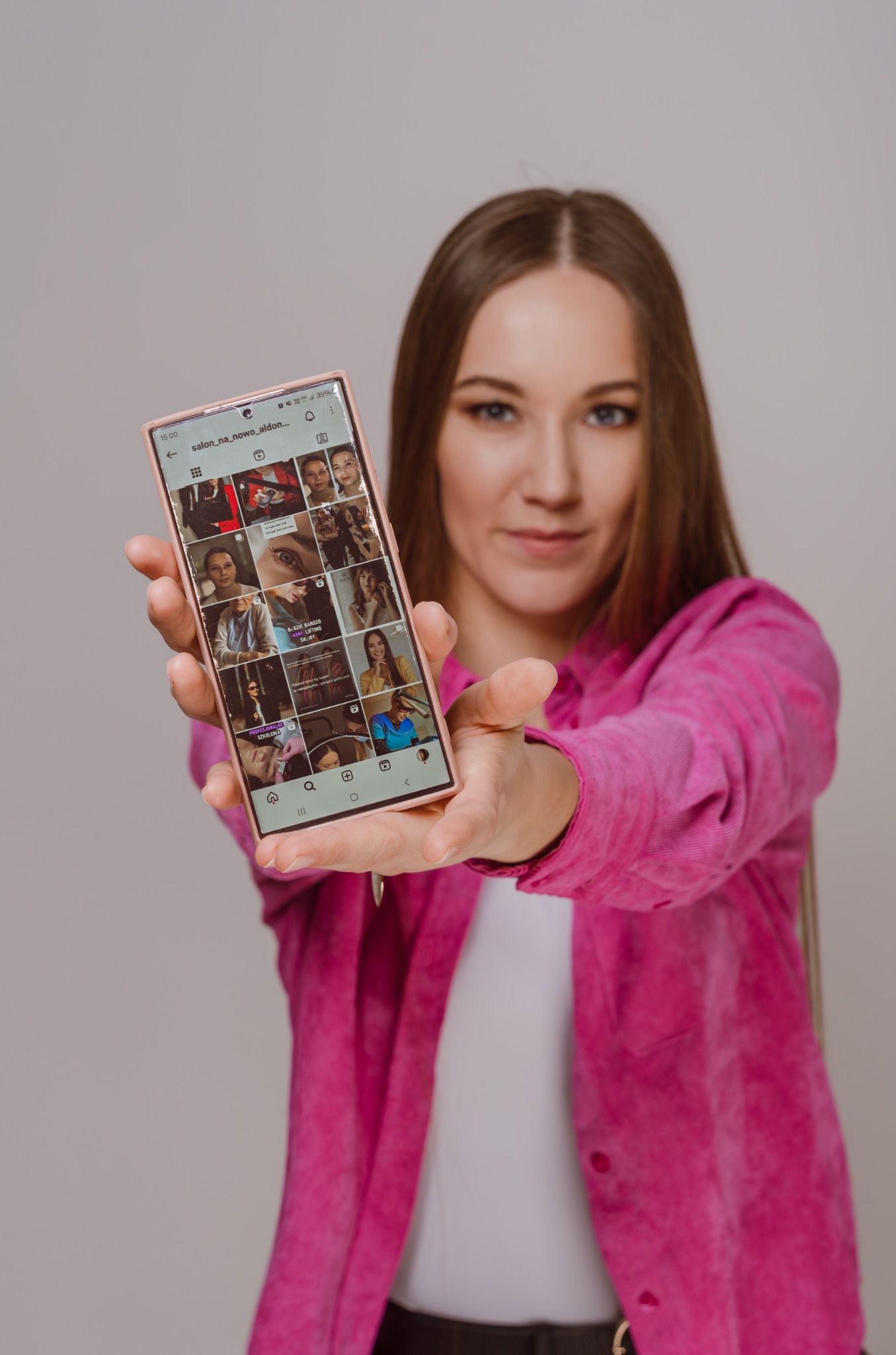 Kobieta w różowej koszuli prezentuje telefon z galerią zdjęć salonu kosmetycznego. Ujęcia makijażu permanentnego, liftingu skóry i szkoleń zawodowych.