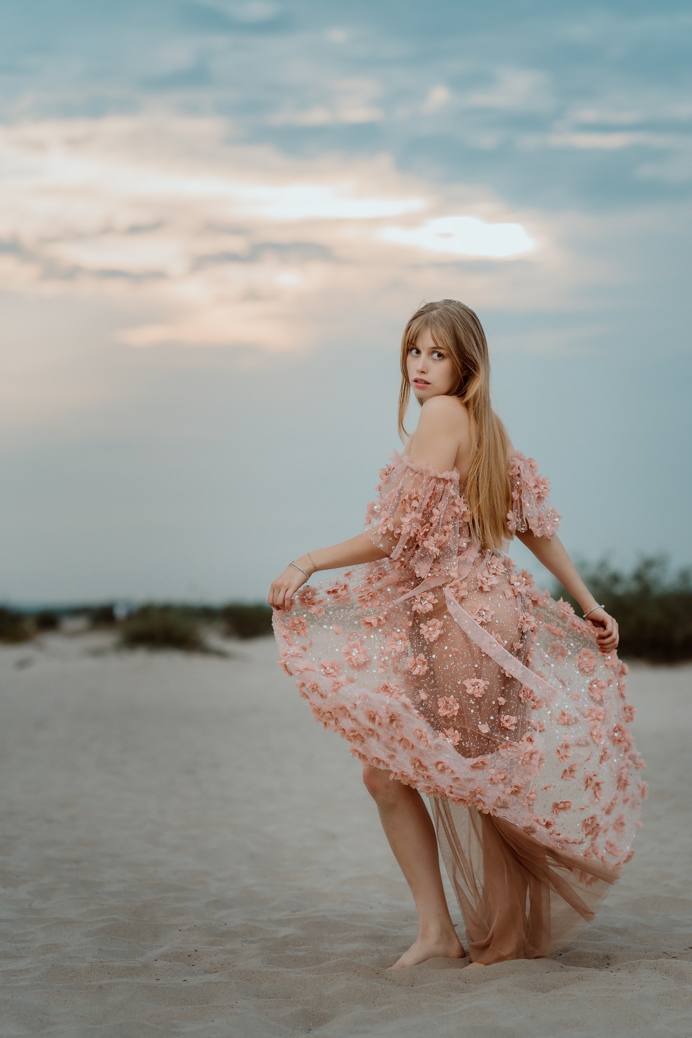 Kobieta w zwiewnej, różowej sukni z kwiatami, odwrócona tyłem, spogląda przez ramię na piaszczystej plaży o zmierzchu. Delikatne światło podkreśla detale sukni.