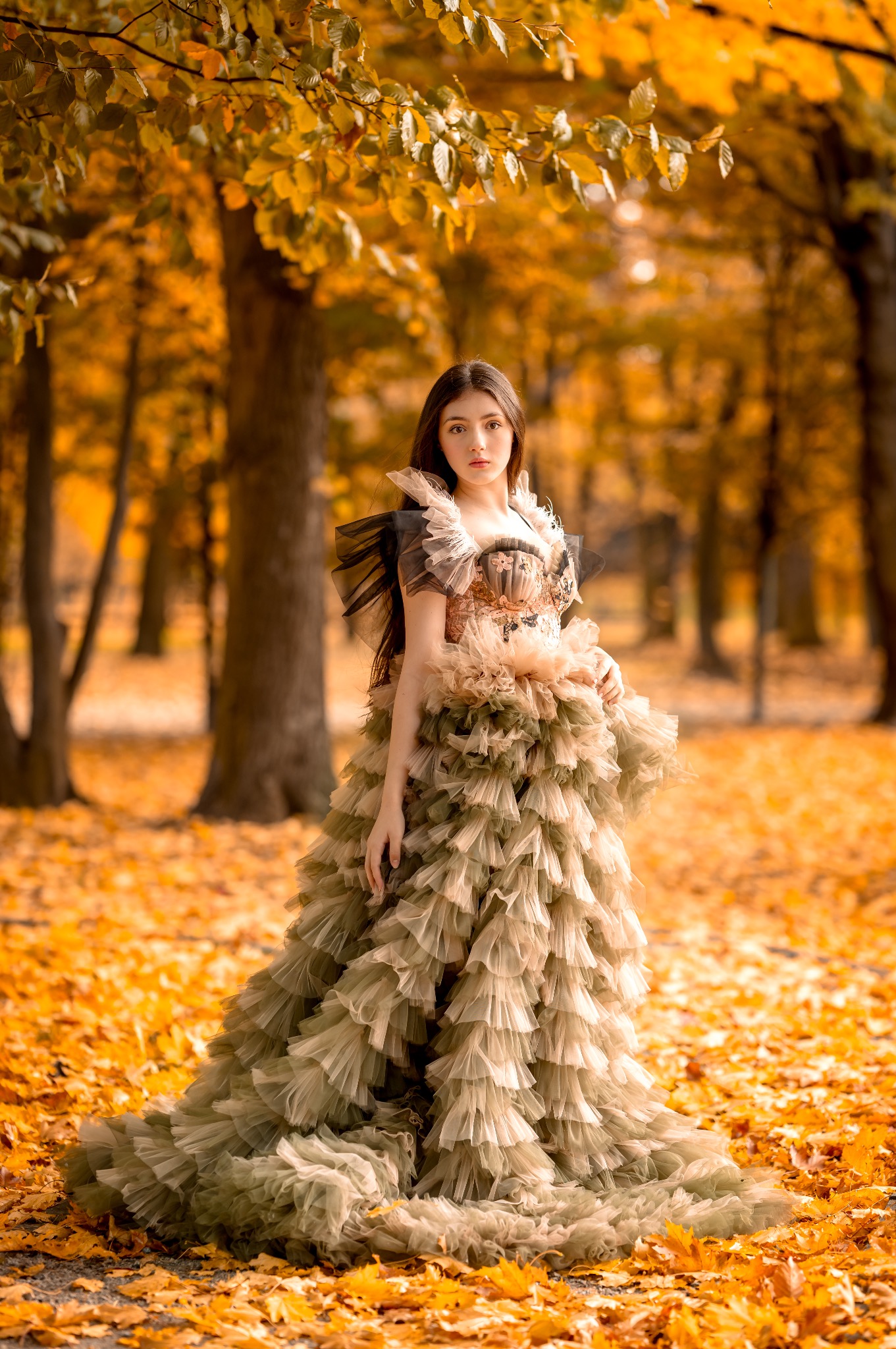 Młoda kobieta w bogatej, warstwowej sukni pozuje w jesiennym lesie. Ciepłe światło podkreśla opadłe liście i detale kreacji. Romantyczna sesja w plenerze.