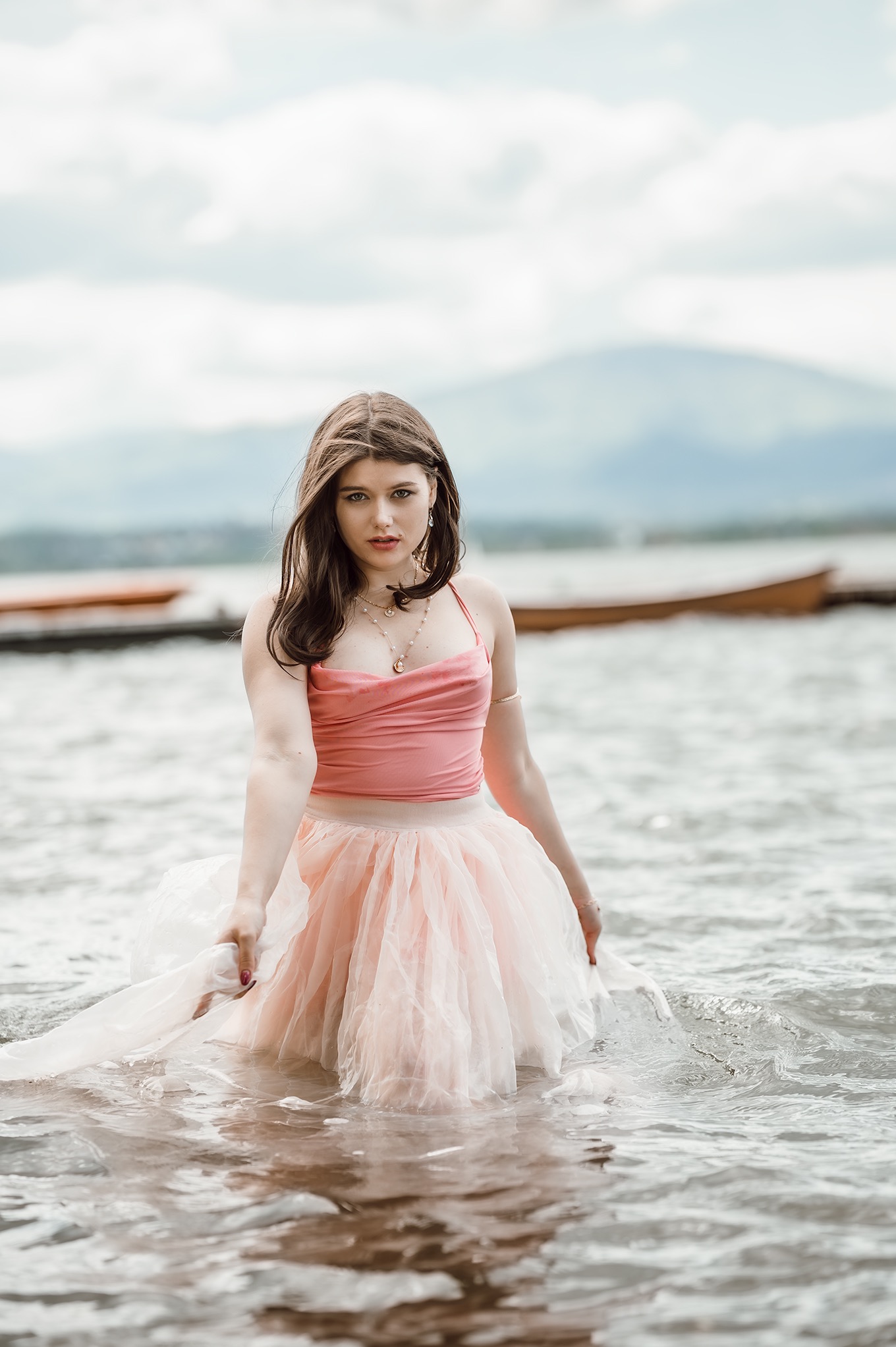 Kobieta w tiulowej sukience w wodzie jeziora, z górami w tle. Ujęcie z dołu podkreśla jej sylwetkę i otoczenie. Delikatne światło i pastelowe kolory tworzą eteryczny klimat.