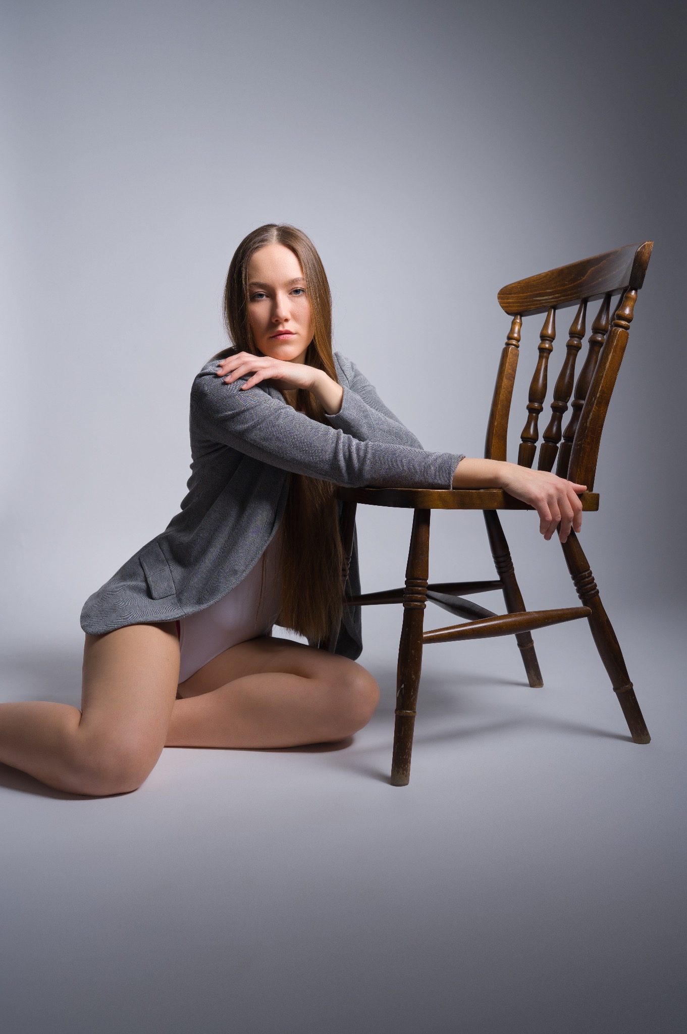 Kobieta w marynarce pozuje siedząc obok krzesła na sesji w Trzebini. Stonowane kolory, profesjonalne oświetlenie, gładkie tło. Długie włosy modelki.