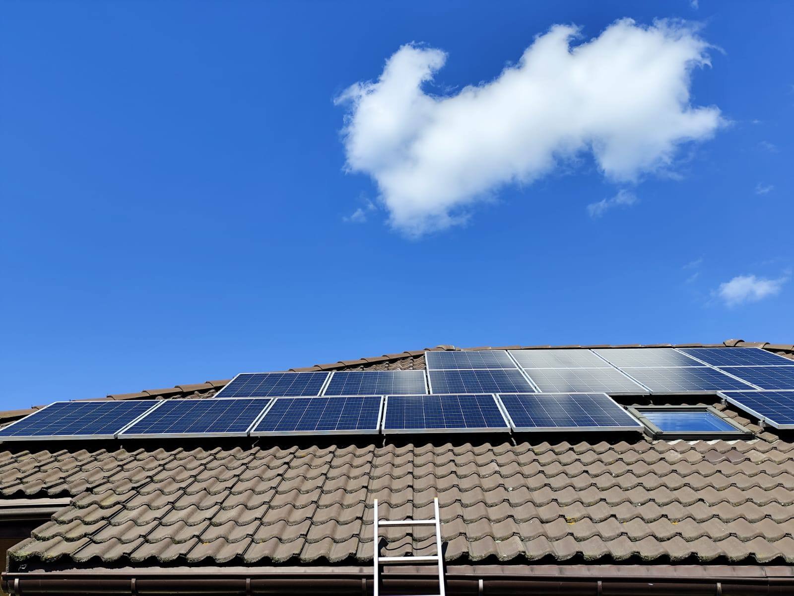 Brązowy dach z panelami słonecznymi w słoneczny dzień. Biała drabina oparta o rynnę. Błękitne niebo z pojedynczą białą chmurą. Widok z dołu.