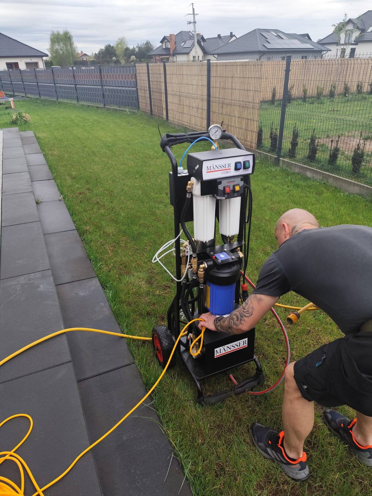 Pracownik z tatuażem obsługuje mobilny system filtracji wody MANSSER na trawniku obok kostki brukowej. Urządzenie z manometrem i filtrami, w tle ogrodzenie i domy.