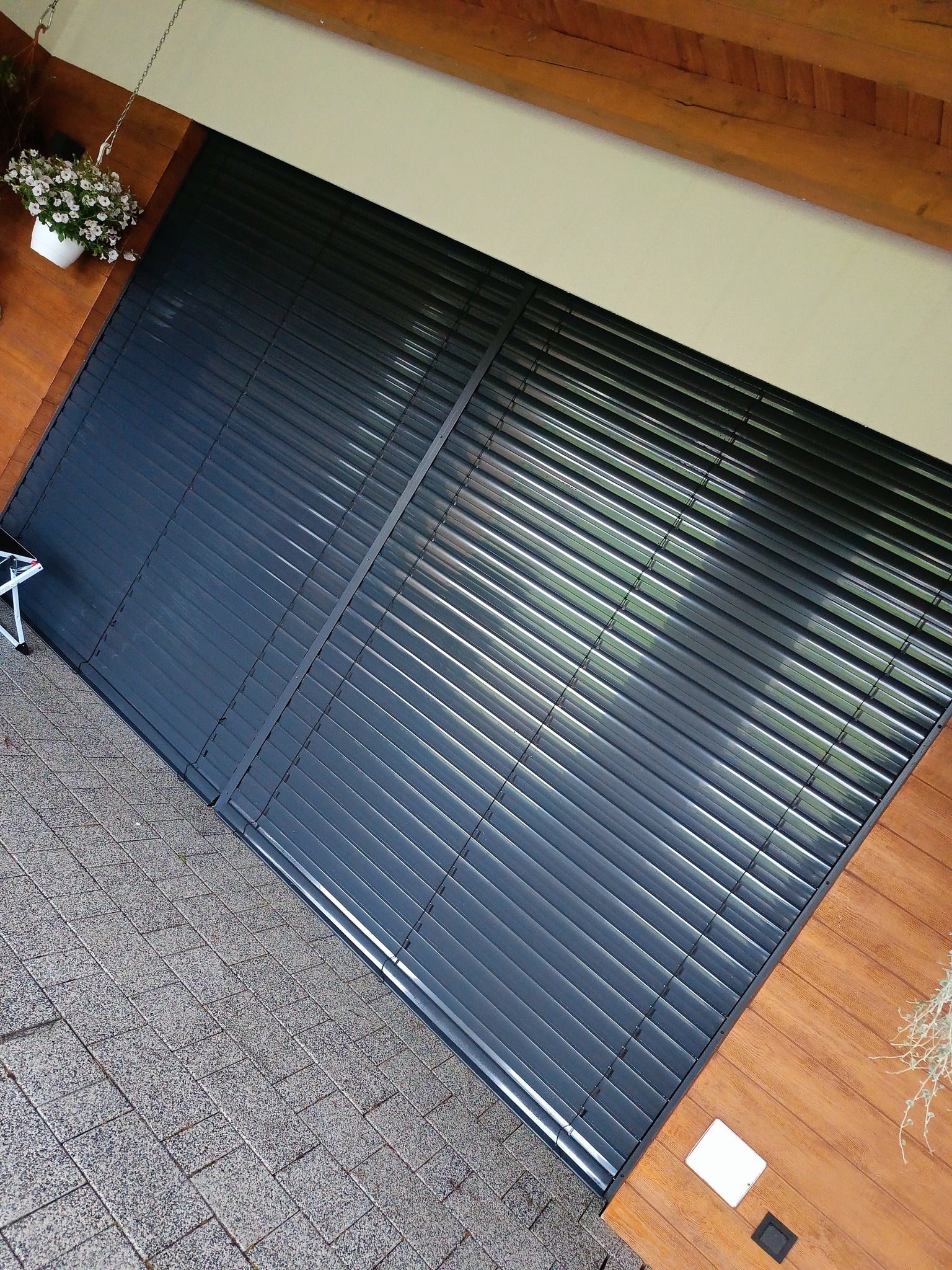 Zewnętrzne, grafitowe żaluzje okienne zamontowane na drewnianej elewacji, widziane z góry. W tle kostka brukowa i donica z kwiatami.