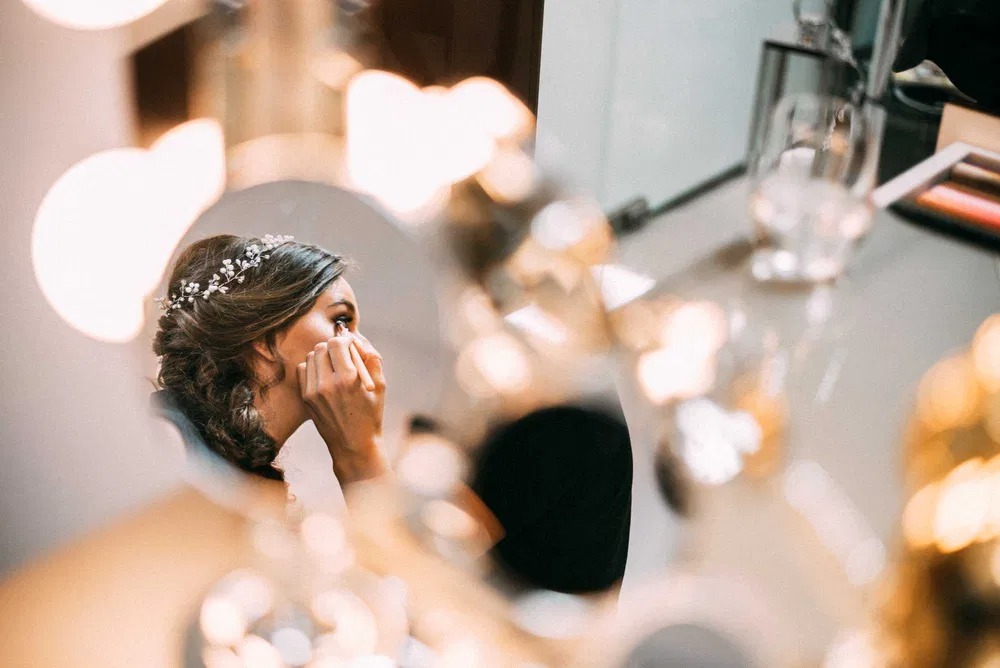 Panna młoda z delikatną ozdobą we włosach maluje rzęsy przed lustrem; w tle kosmetyki i szklanka wody.