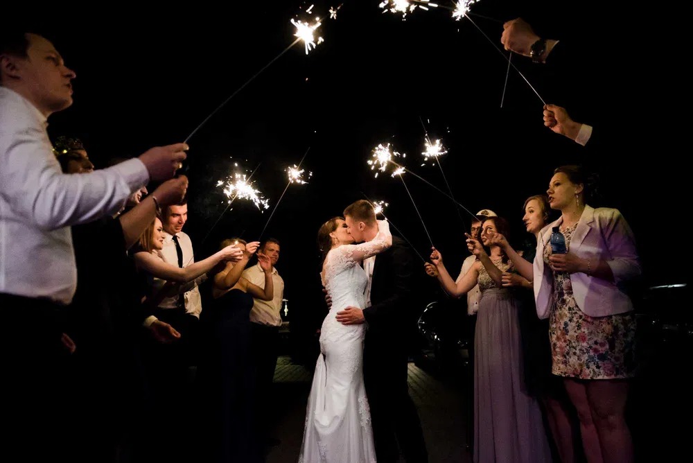 Pocałunek pary młodej otoczonej gośćmi z zimnymi ogniami w ciemności.