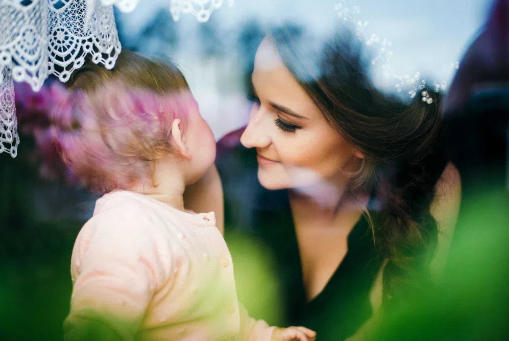 Portret matki spoglądającej z czułością na małe dziecko, widoczne przez szybę z odbiciami i elementami dekoracyjnymi.