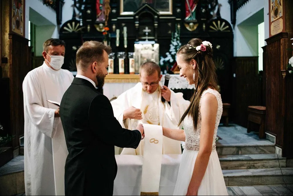 Para młoda podczas ceremonii ślubnej w kościele, ksiądz w maseczce błogosławi obrączki na stułę.