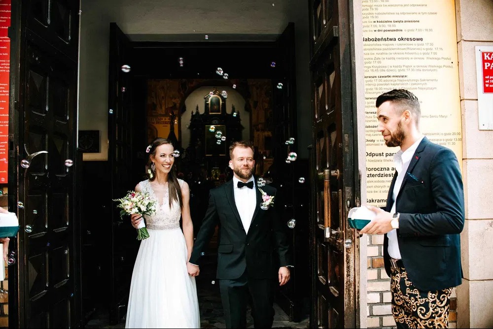 Para młoda wychodzi z kościoła obsypana bańkami mydlanymi, mężczyzna w ekstrawaganckich spodniach trzyma pojemnik na płyn do robienia baniek.