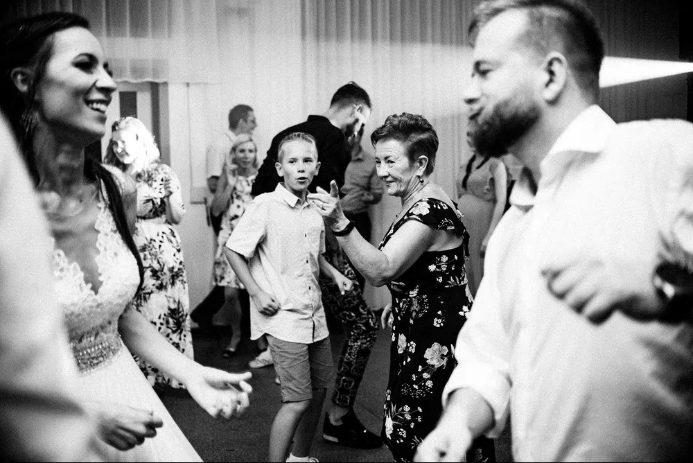 Czarno-białe zdjęcie z wesela: uśmiechnięta panna młoda w koronkowej sukni, energicznie tańczący chłopiec z otwartymi ustami, kobieta w kwiecistej sukience wskazująca palcem, oraz mężczyzna z brodą...