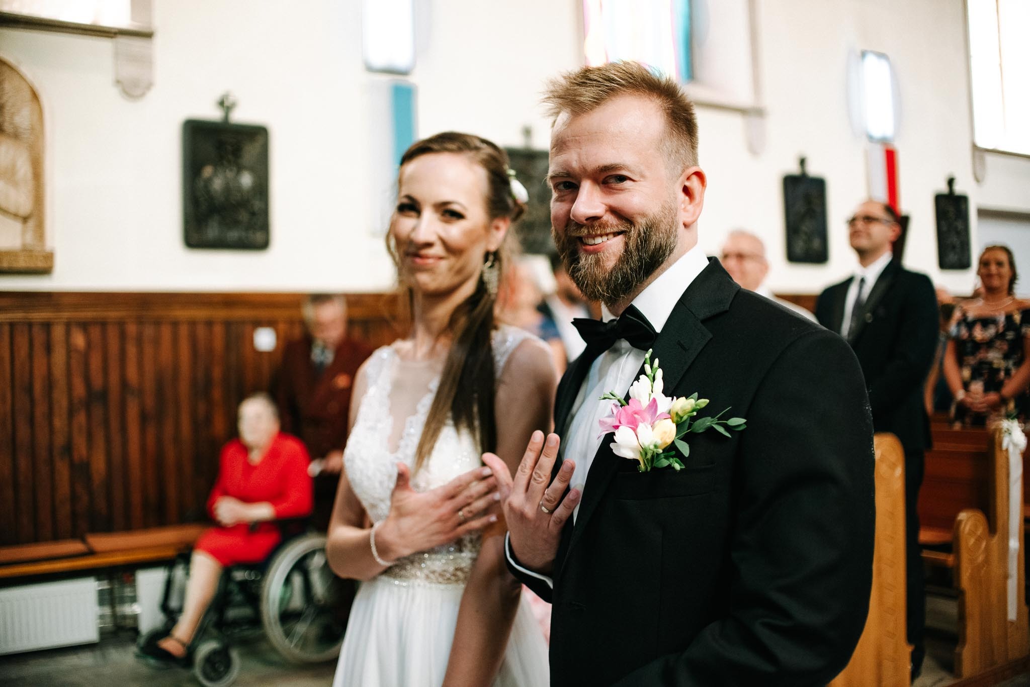 Uśmiechnięta para młoda w kościele, panna młoda w koronkowej sukni, pan młody w czarnym smokingu z butonierką, obrączki na palcach, goście w tle.