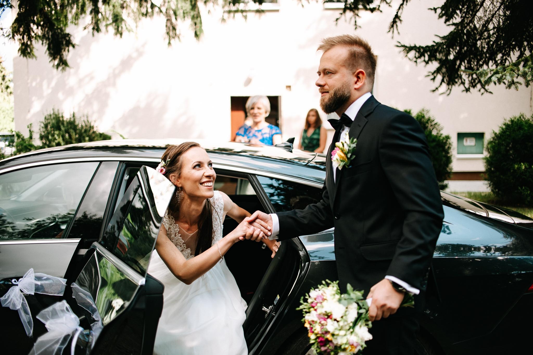 Uśmiechnięta panna młoda wysiada z czarnego samochodu, trzymając za rękę pana młodego w garniturze z butonierką; w tle budynek i obserwujące osoby.