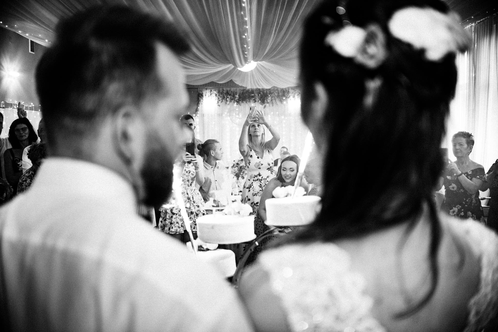 Czarno-białe ujęcie z wesela, goście robią zdjęcia telefonami, w tle dekoracje i tort weselny.