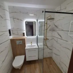 Nowoczesna łazienka z marmurowymi płytkami ściennymi, drewnianą podłogą, podświetlanym lustrem nad białą szafką z umywalką i kabiną prysznicową ze złotą armaturą.