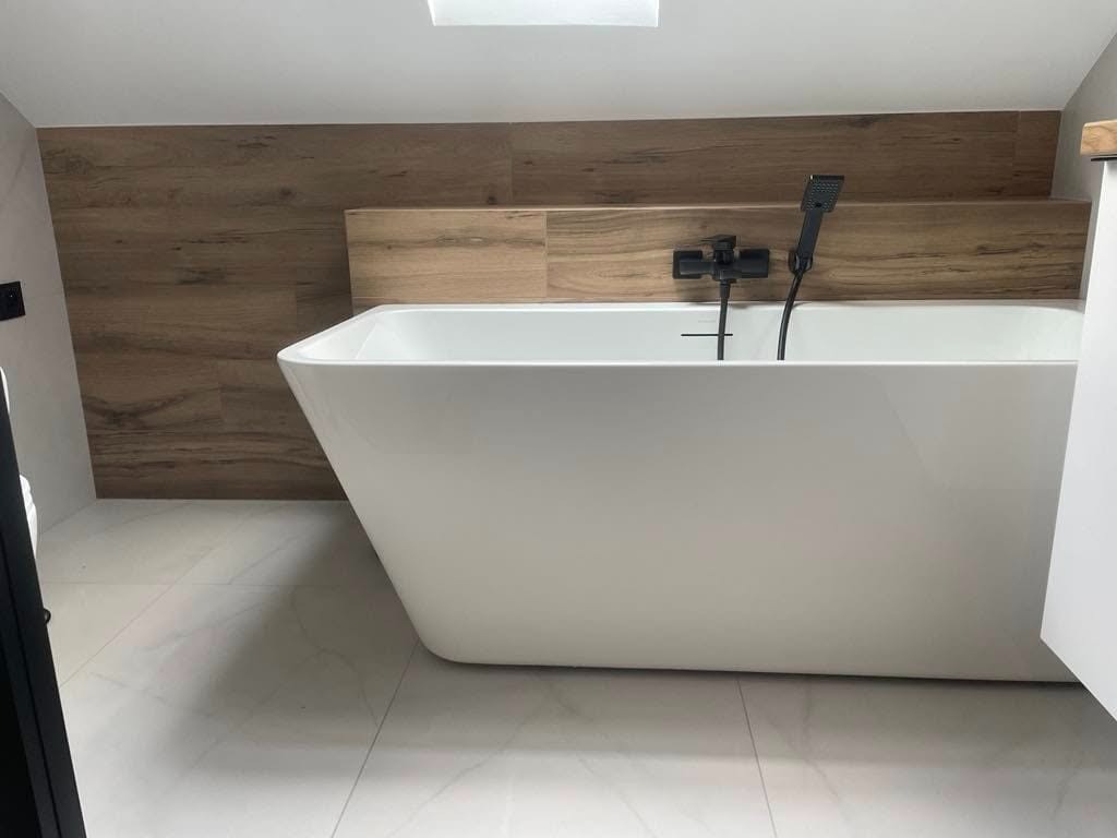 Nowoczesna, wolnostojąca wanna z czarną armaturą i drewnopodobną okładziną ścienną w łazience wykończonej białymi płytkami.