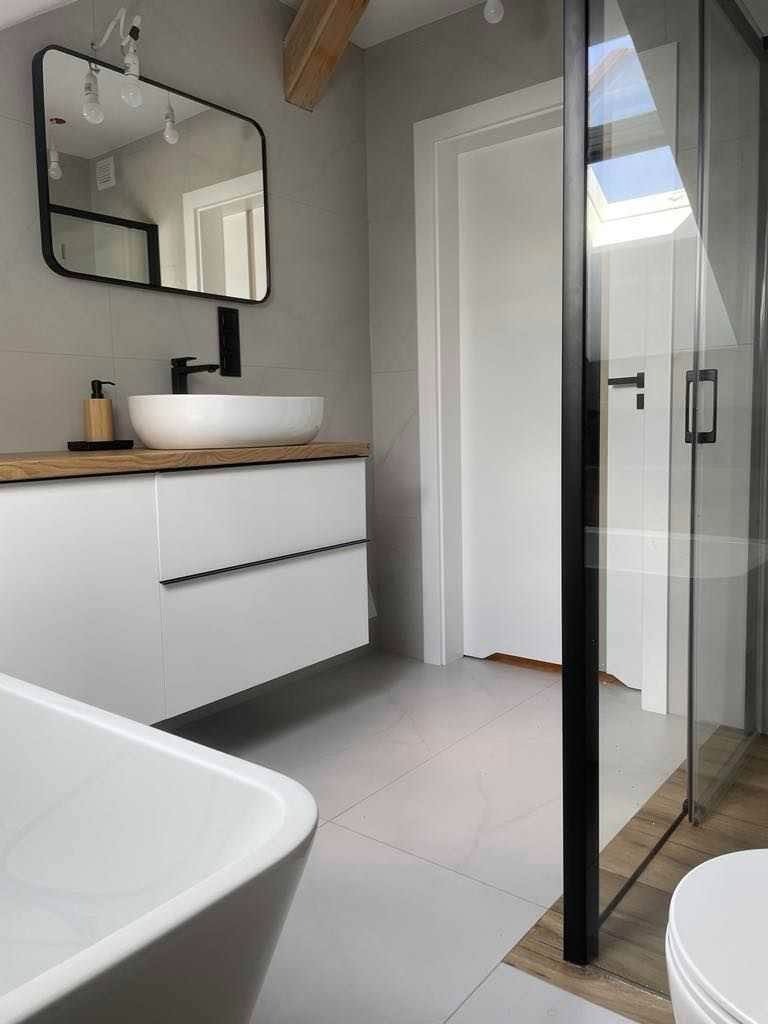 Nowoczesna łazienka z umywalką nablatową, czarną armaturą, lustrem w czarnej ramie, białą szafką pod umywalką z czarnymi uchwytami, kabiną prysznicową ze szklanymi drzwiami i drewnianym akcentem...