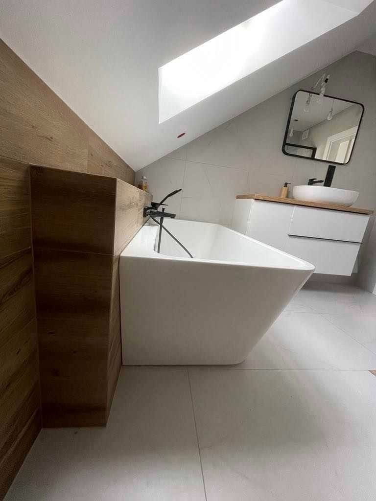 Nowoczesna łazienka na poddaszu z białą, prostokątną wanną, drewnianą zabudową, umywalką nablatową i lustrem w czarnej ramie, doświetlona oknem dachowym.