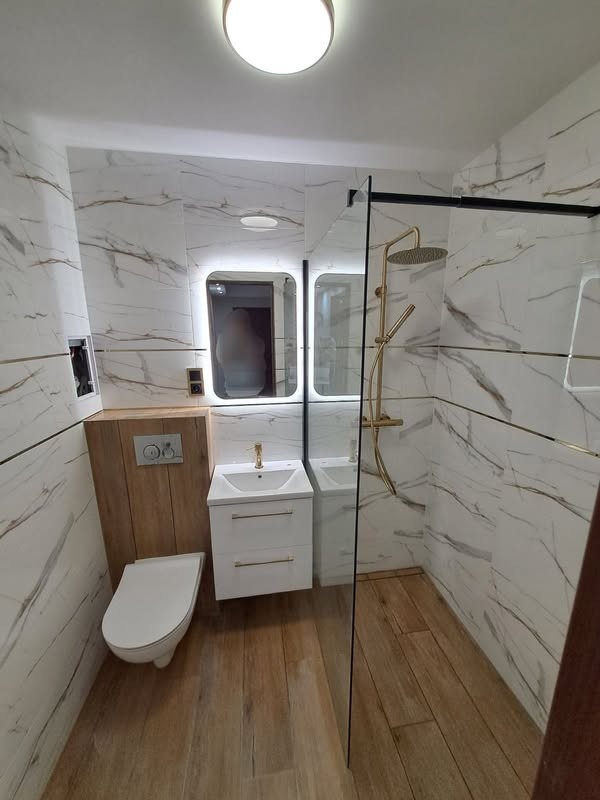Nowoczesna łazienka z marmurowymi płytkami ściennymi, drewnianą podłogą, podświetlanym lustrem nad białą szafką z umywalką i kabiną prysznicową ze złotą armaturą.