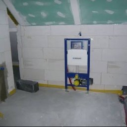 HYDROMAT - Łazienka w stanie deweloperskim z zamontowanym niebieskim stelażem podtynkowym Geberit do WC, widoczne białe ściany z bloczków, zielony sufit z płyt GK i żółta taśma izolacyjna na posadzce.