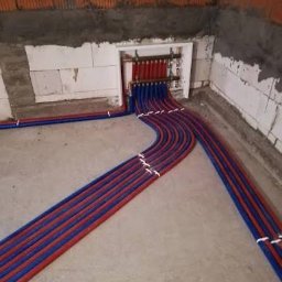 HYDROMAT - Starannie ułożone czerwone i niebieskie rury instalacji grzewczej na betonowej posadzce, prowadzące do rozdzielacza wnękowego. Surowe ściany z cegły i bloczków.