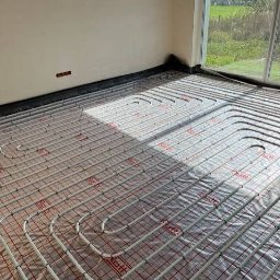 HYDROMAT - Instalacja ogrzewania podłogowego z białymi rurami ułożonymi na czerwonej siatce izolacyjnej w jasnym pomieszczeniu. Słońce rzuca cienie na podłogę, a przez duże okno widać zielony krajobraz.
