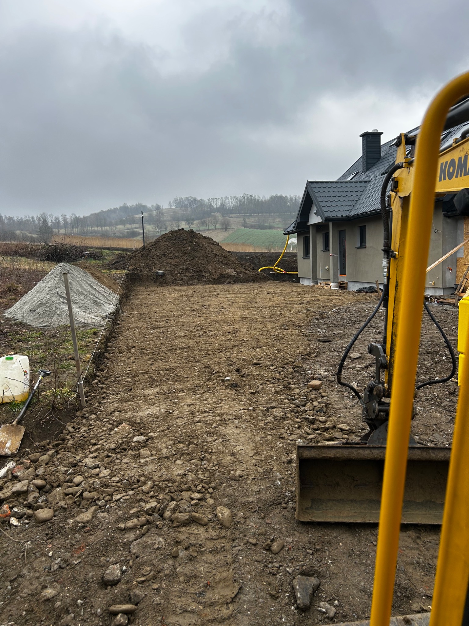 Przygotowanie terenu pod budowę domu jednorodzinnego: wyrównany grunt, pryzmy piasku i ziemi, widoczny fragment koparki Komatsu, w tle zalesione wzgórza pod pochmurnym niebem.
