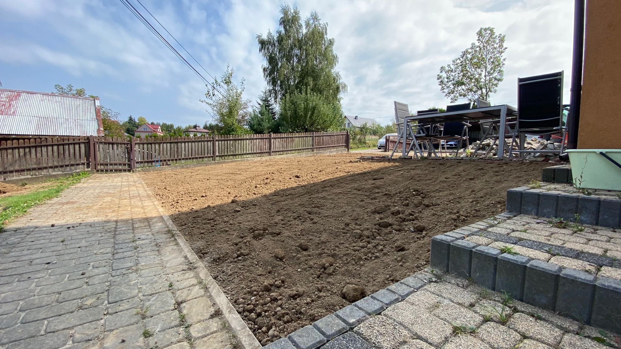 Przygotowany teren pod trawnik lub nasadzenia, widoczne wyrównane podłoże z ziemi, obok kostka brukowa i schody, w tle ogrodzenie posesji i drzewa.
