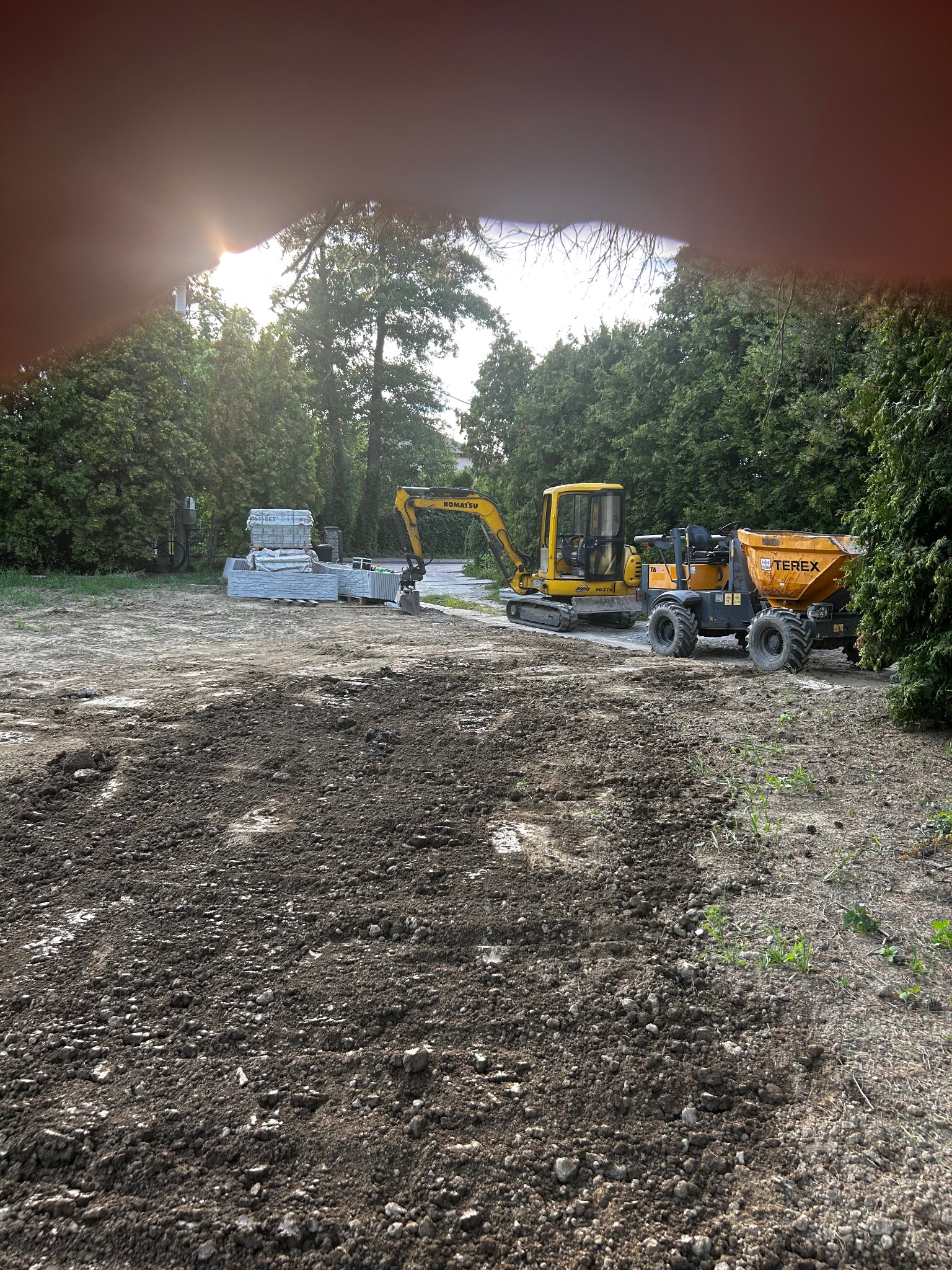 Żółta koparka i wywrotka Terex na placu budowy, w tle palety z materiałami budowlanymi. Teren częściowo przygotowany, otoczenie drzewa.