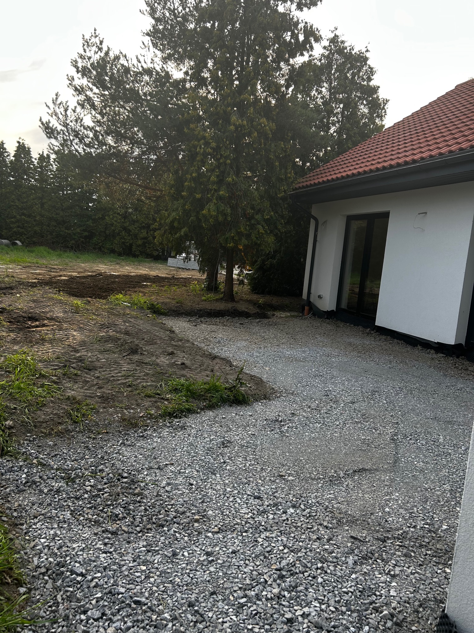 Działka po niwelacji z fragmentem utwardzonej drogi z grysu, obok nowoczesny dom z czerwonym dachem. Widoczne ślady po pracy maszyn.