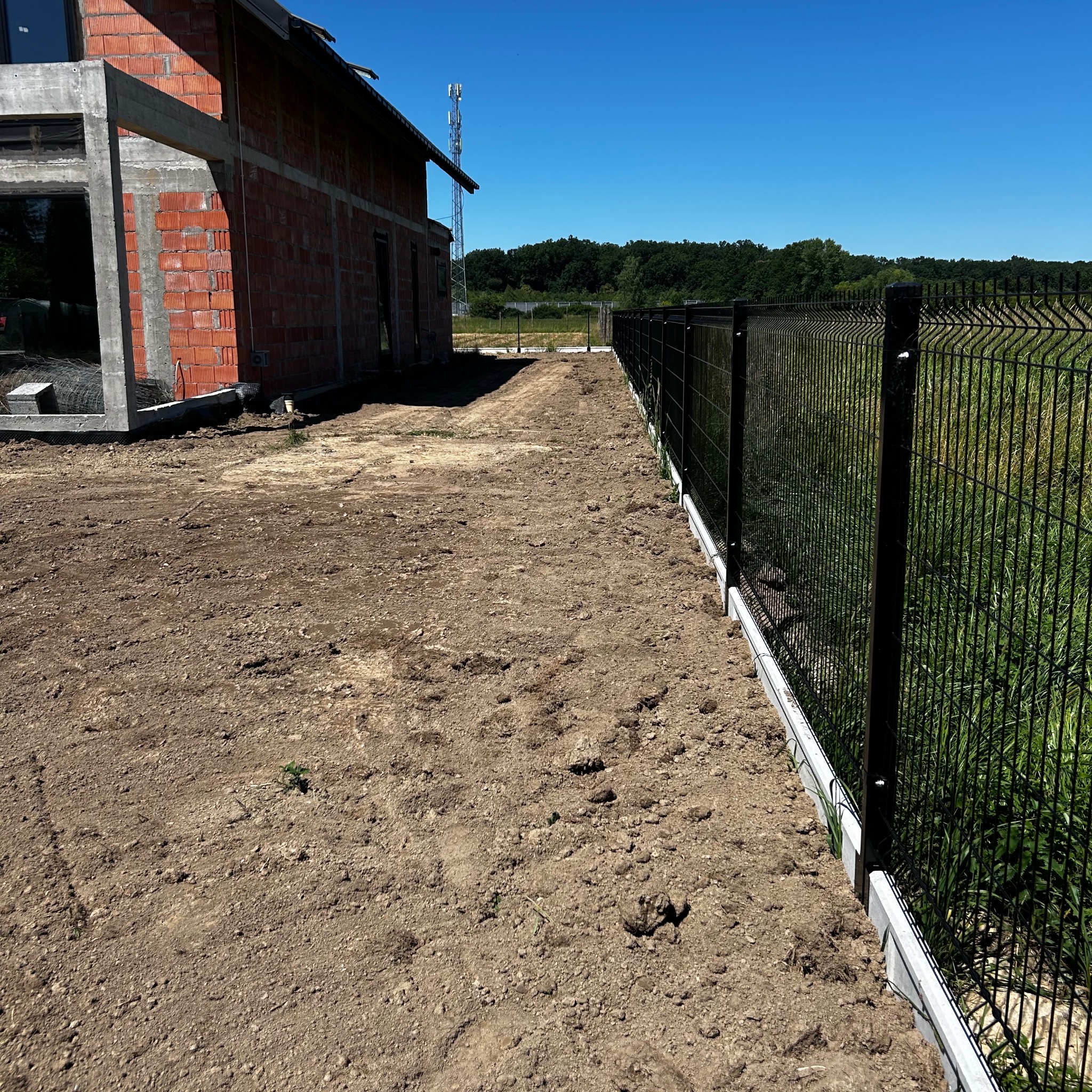 Nowo postawione, czarne ogrodzenie panelowe z podmurówką na wyrównanym terenie obok budynku w stanie surowym otwartym, w tle zieleń i wieża telekomunikacyjna.