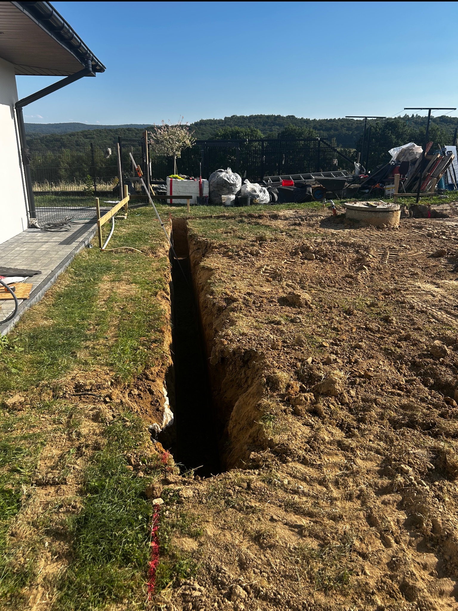 Wąski wykop w ziemi, prawdopodobnie pod instalację, z widocznym fragmentem budynku i elementami ogrodzenia w tle. Teren przygotowany pod dalsze prace.