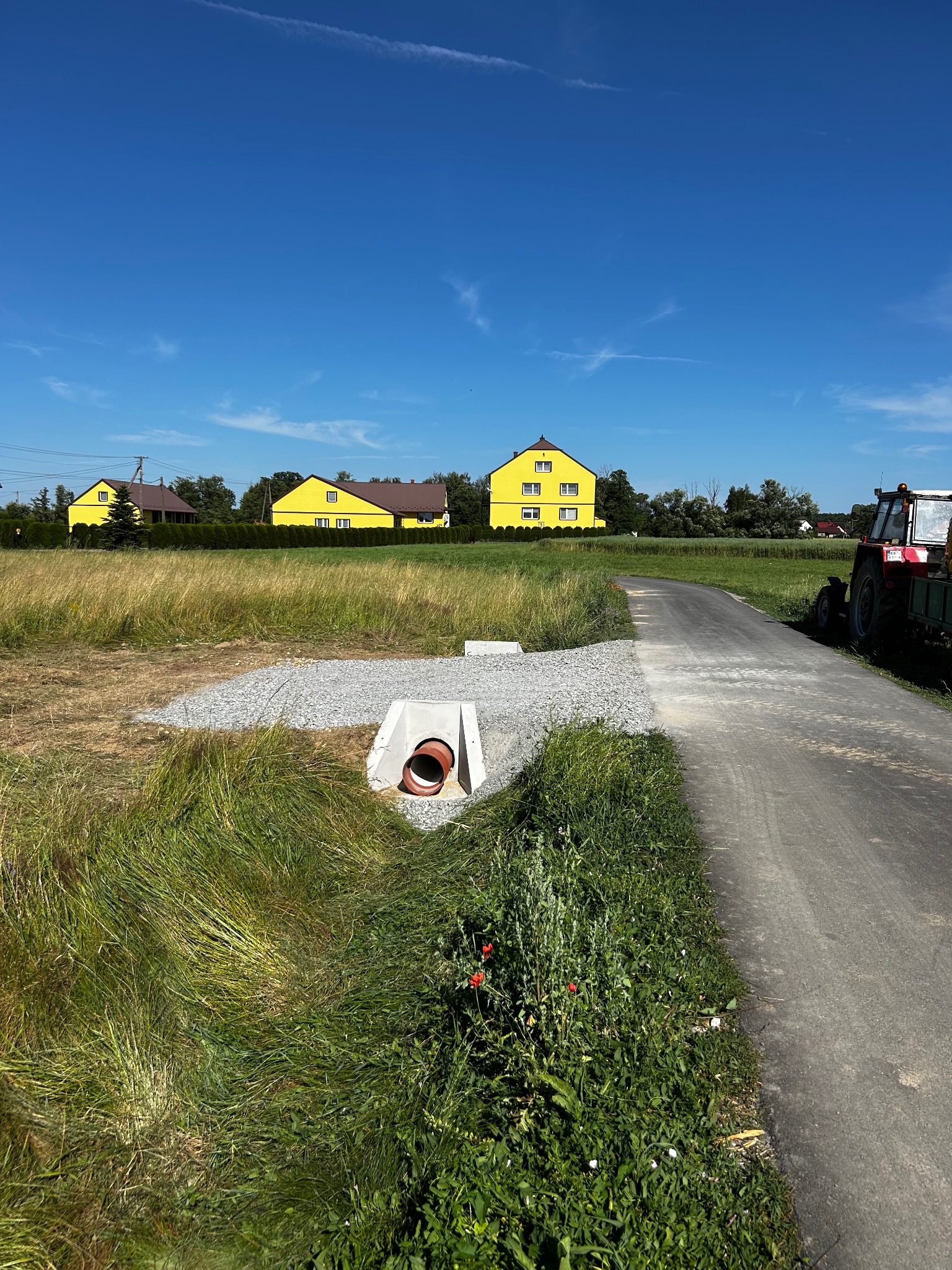 Betonowy przepust z rurą na skraju drogi i pola, w tle żółte budynki i błękitne niebo. Po prawej traktor z przyczepą. Widok na system odwadniający.