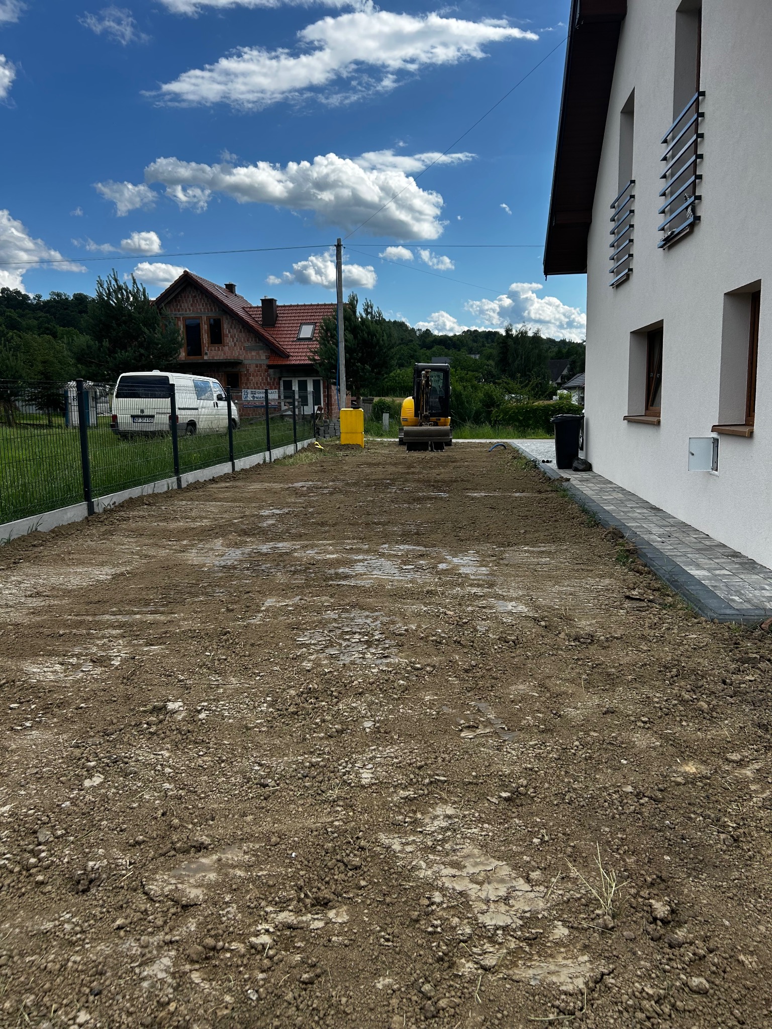 Przygotowany teren pod budowę drogi dojazdowej z widocznym minikoparką i fragmentami kostki brukowej. Dom w tle. Błękitne niebo z chmurami.