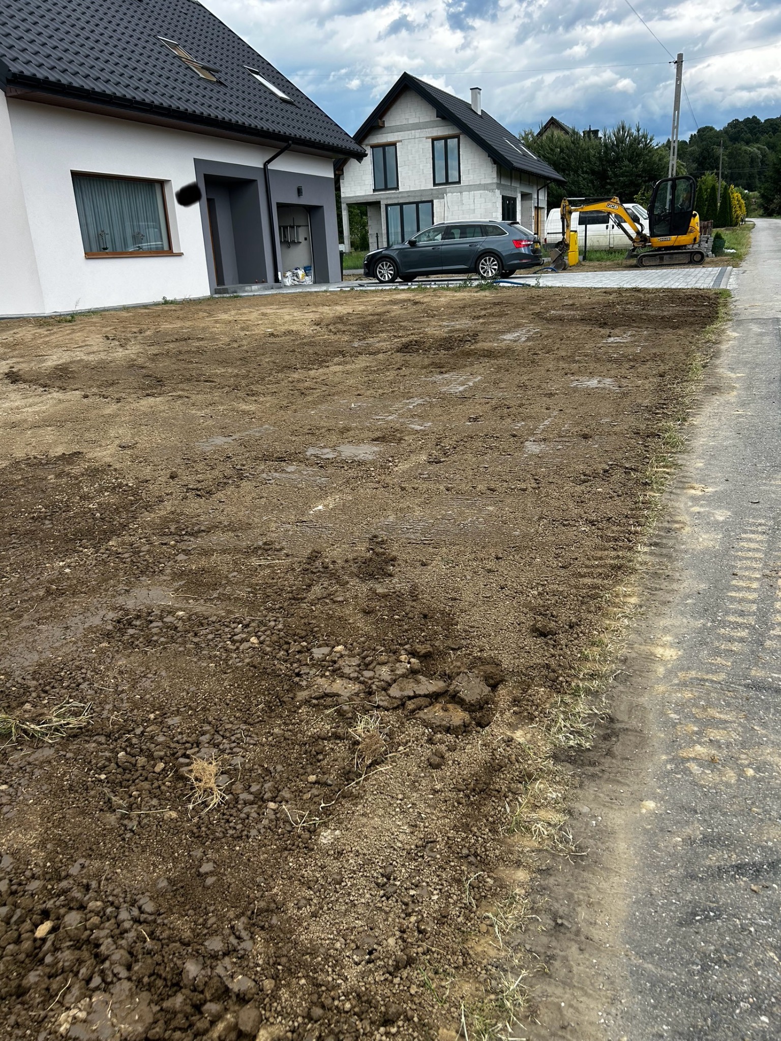 Przygotowany teren pod budowę domu z widocznym małym sprzętem budowlanym. Świeżo wyrównana ziemia, gotowa pod dalsze prace. Dom w tle.